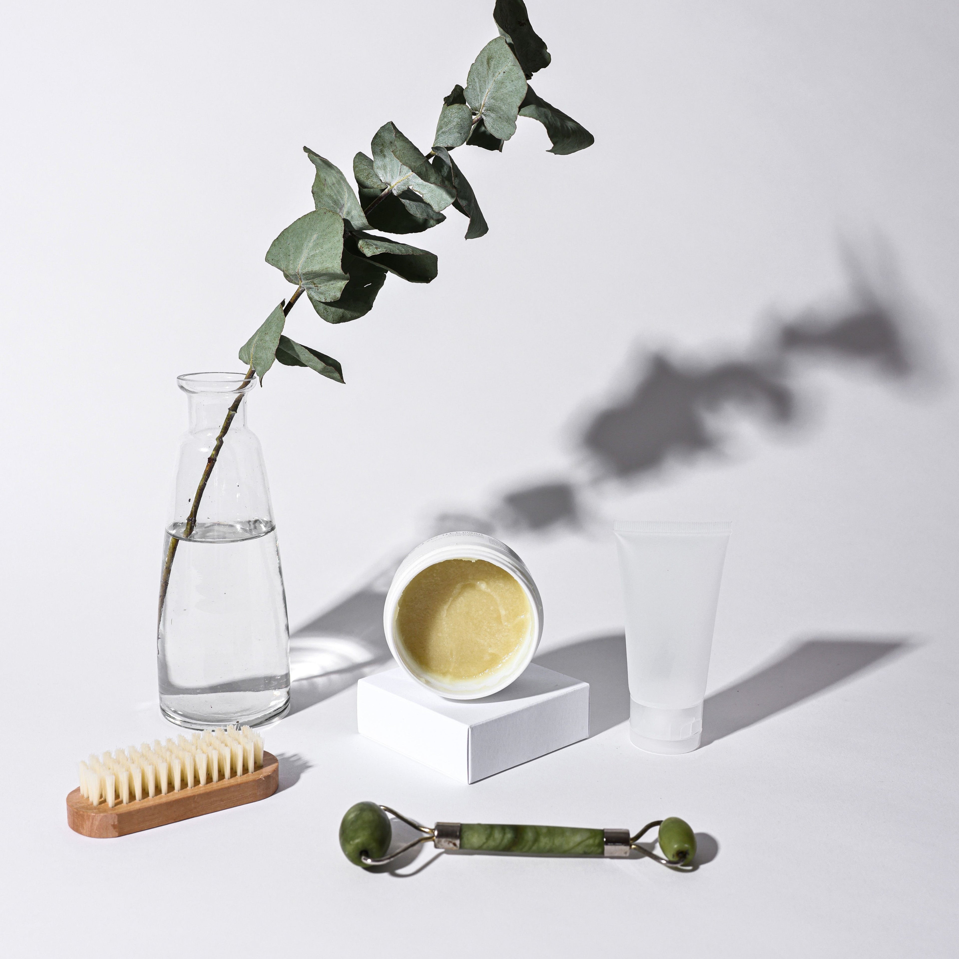 A collection of personal items on a white backgroiund, including a bottle of lotion, a wood scrubbie, and a Green Stone face roller