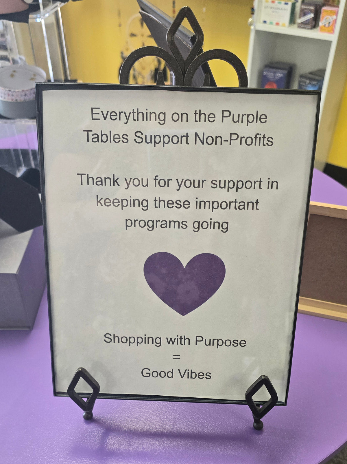 A printed sign on a black stand with a purple heart, reading: 'Everything on the Purple Tables Support Non-Profits' and 'Shopping with Purpose = Good Vibes,' displayed on a purple table in a store setting.