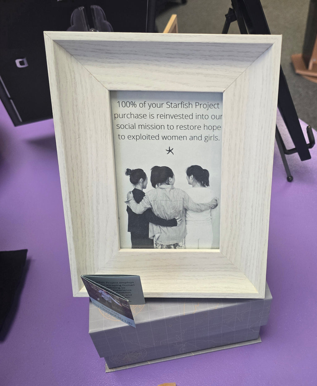 A light wood picture frame displayed on a gray stand against a purple surface, with a printed message and a black-and-white photo inside the frame.