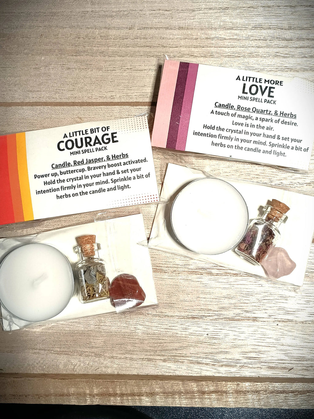 Two small spell packs labeled 'A Little Bit of Courage' and 'A Little More Love' with candles and crystals on a wooden surface.