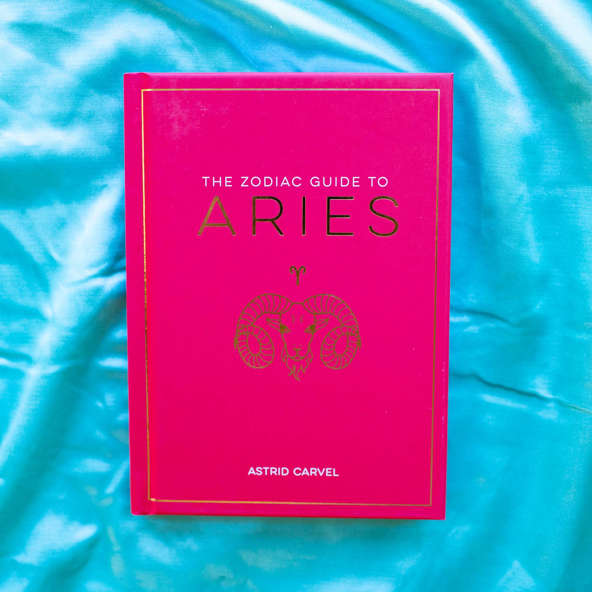 Pink book titled 'The Zodiac Guide to Aries' by Astrid Carvel on a blue textured background