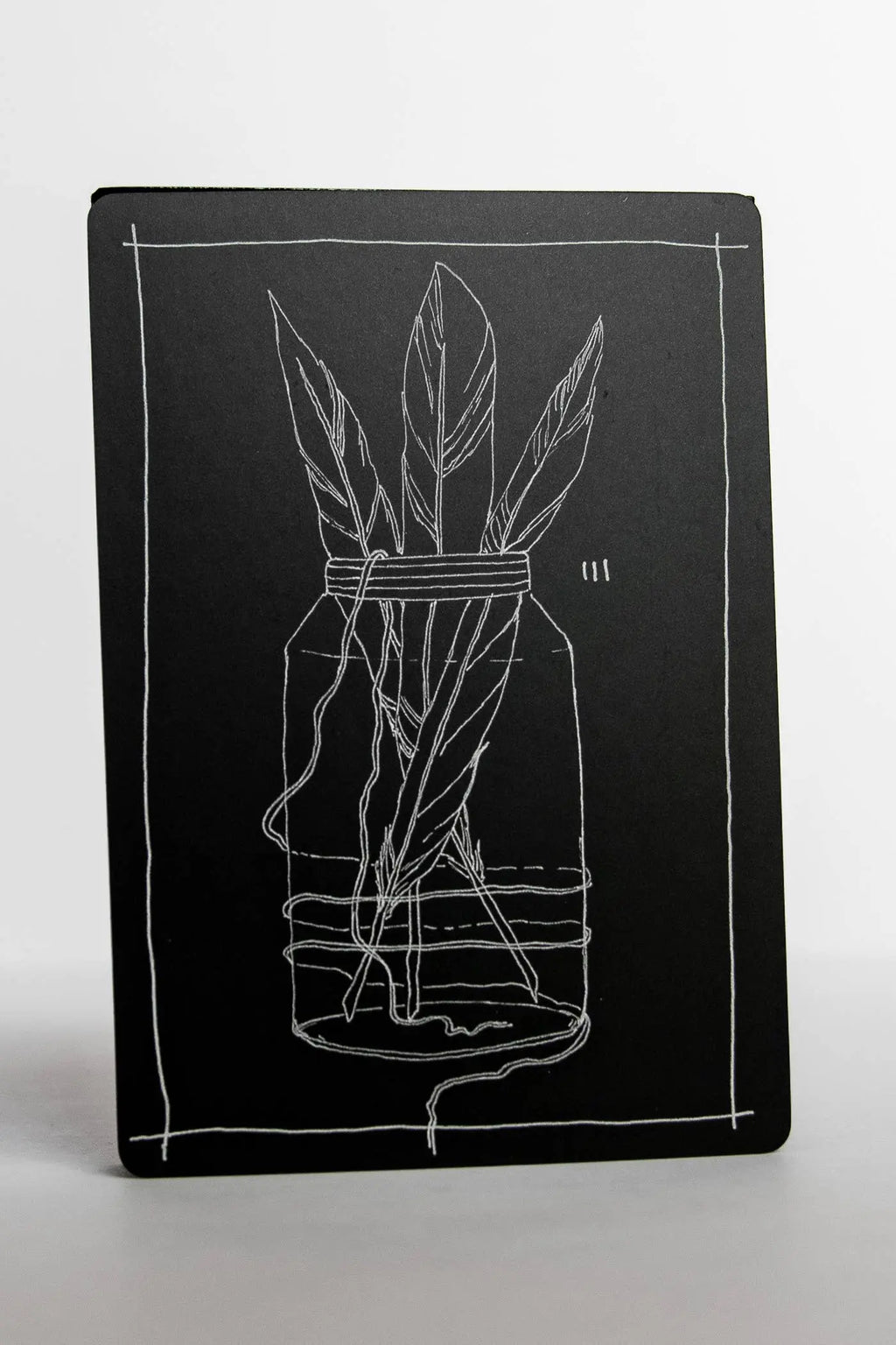 Black notebook with white line drawing of a jar with feathers on a white background