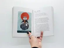 An open book held by a hand, showing a printed page with text on the right and a colorful illustration on the left. The illustration depicts a wolf in front of a red moon against a light blue background.