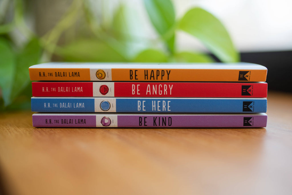 Stack of books titled 'Be Happy', 'Be Angry', 'Be Here', and 'Be Kind' by the Dalai Lama on a wooden surface with blurred greenery in the background.