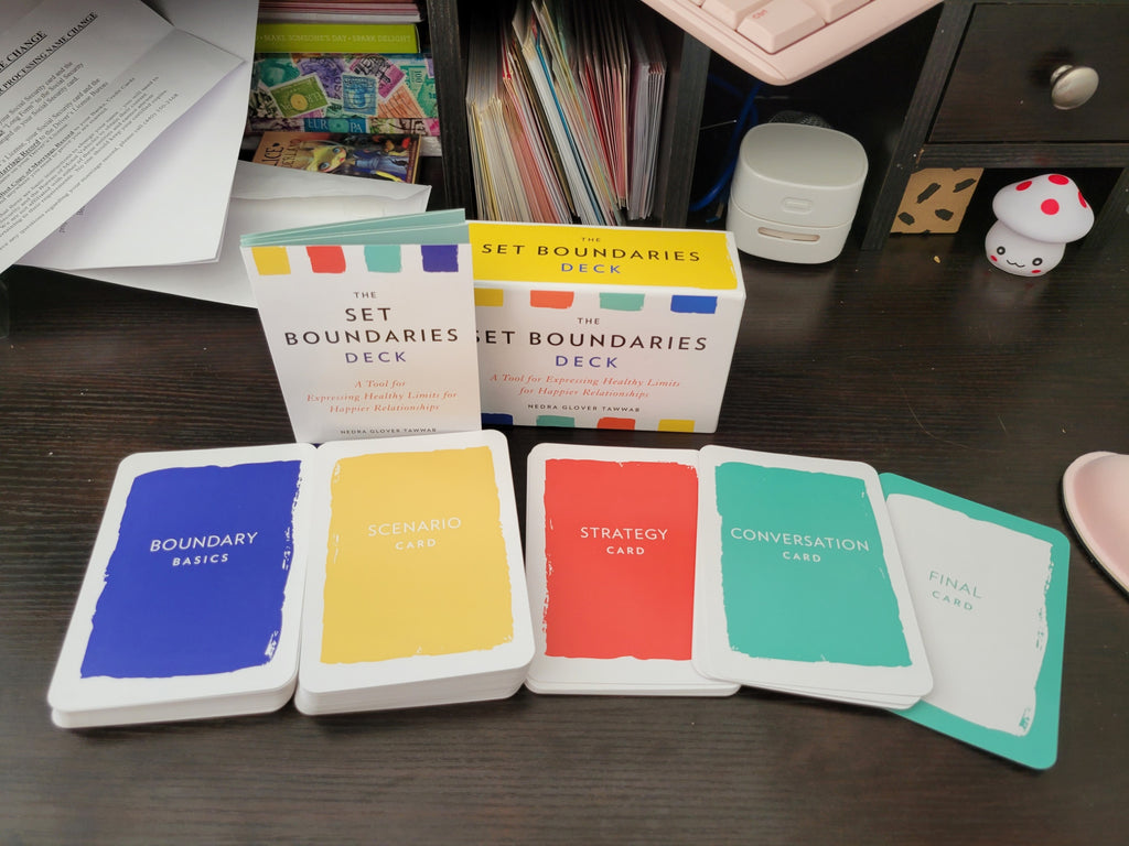 A set of colorful card decks and individual cards from 'The Set Boundaries Deck' arranged on a dark wooden desk, with papers and other desk items in the background.
